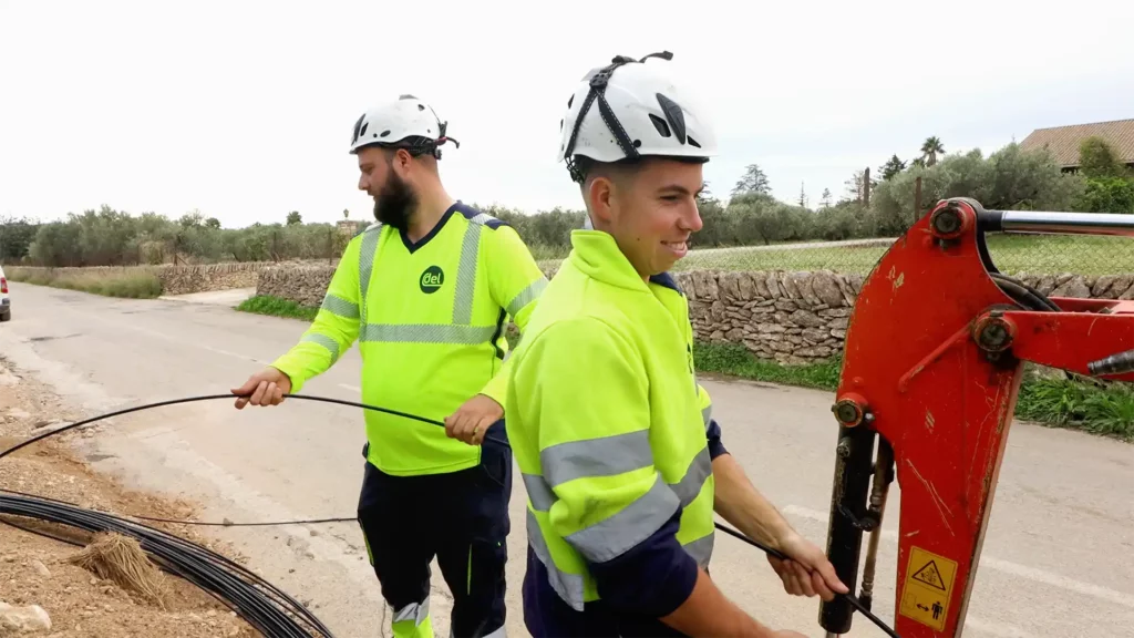 Dos treballadors de l'empresa Delinternet, amb roba d'alta visibilitat i cascos de seguretat, desplegant cable de fibra òptica al costat d'una carretera a la població dels Reguers, utilitzant maquinària i bobines de cable.